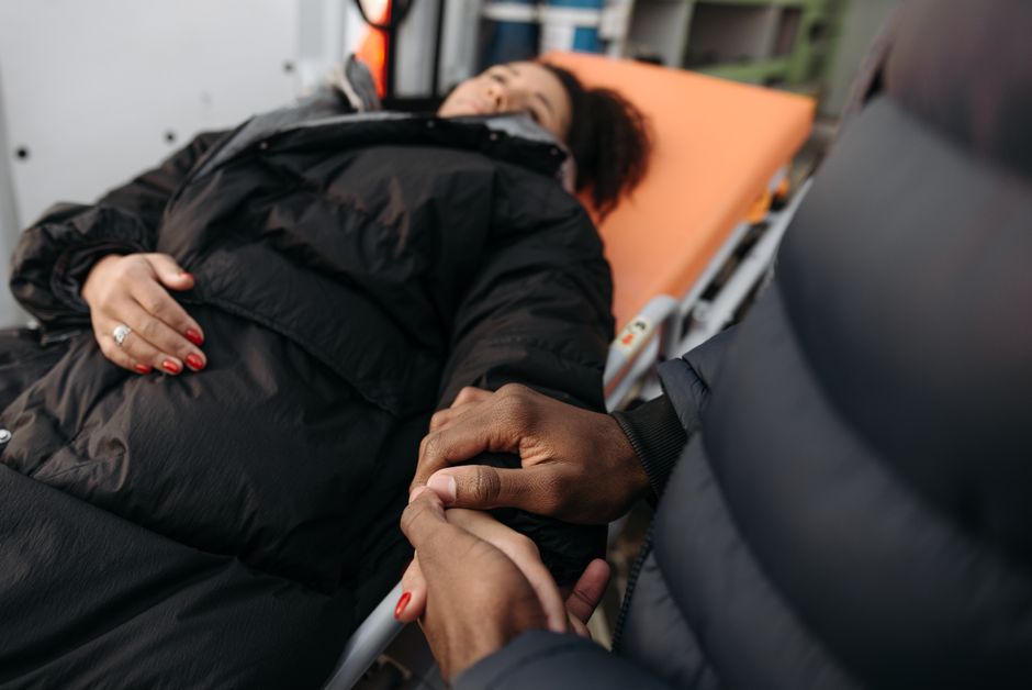 A woman lying on a hospital bed with an orange mattress, dressed in a black jacket with visible red nail polish on her hand resting on her abdomen. An individual with dark skin, wearing a black jacket