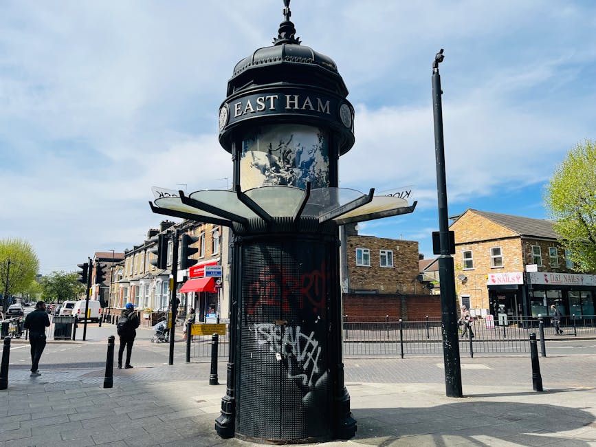 A large, cylindrical urban information kiosk with a rounded, dome-shaped top displaying the text 'EAST HAM' on a sign. The kiosk is covered with graffiti and is situated on a paved pavement at a busy intersection in Tooting, London. In the background, there are several pedestrians walking across the crosswalk, residential buildings with brick facades, and a small local shop with a red and white sign. The sky is partly cloudy with patches of blue, and surrounding trees have fresh green leaves. The area appears to be a lively part of the neighbourhood, with nearby light poles and street furniture, providing context for the urban environment. This setting may relate to house removals or local moving services in the Tooting area, with Tooting Movers operating in this vicinity for furniture transport and home relocations.
